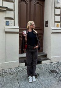 Kvinna med blont hår, mörk jacka hängande över axlarna, svart topp, bruna byxor, vita sneakers, hållande en kaffekopp, stående framför en brun dörr.
