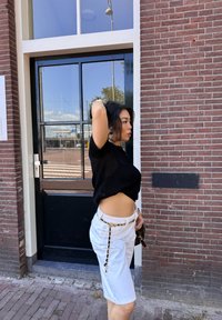 Zwarte gebreide crop top, witte high-waisted shorts met een patroonriem, staand tegen een bakstenen muur en een grote glazen deur.