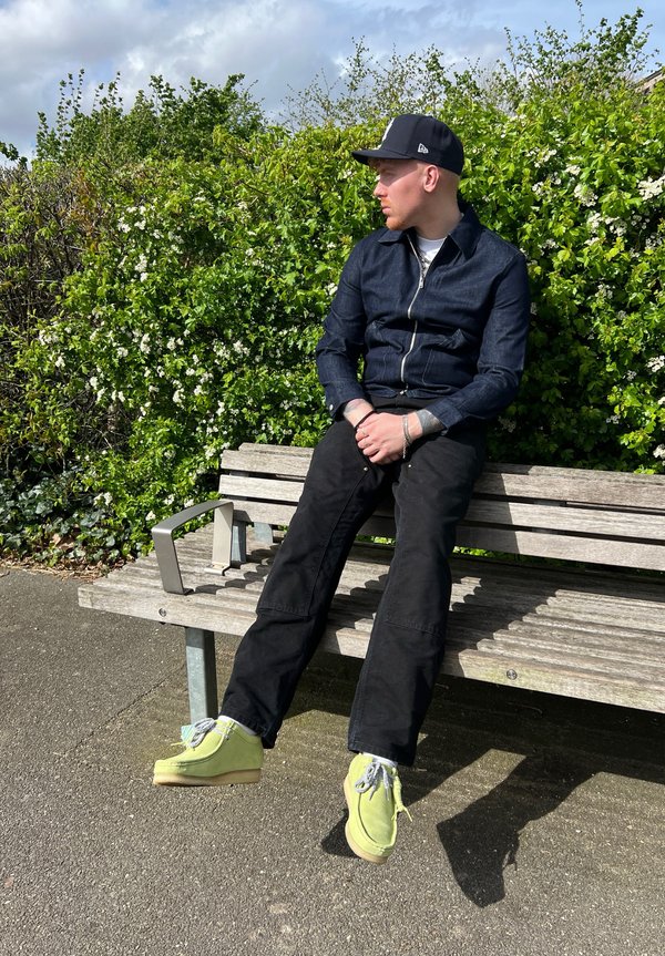 Chaqueta de mezclilla negra con cremallera, pantalones negros y zapatos de ante verde lima sobre un banco de madera, con un fondo de follaje verde y flores blancas.