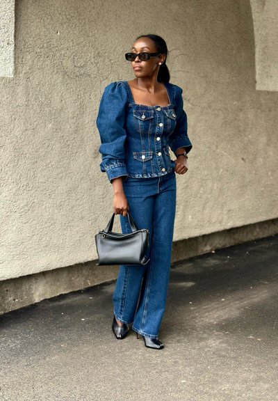 Mujer con chaqueta de mezclilla de mangas abullonadas y jeans, sosteniendo un bolso negro, con tacones negros puntiagudos y gafas de sol oscuras, de pie al aire libre.