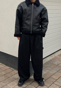 Veste en cuir noire avec col en fourrure, fermeture éclair, poignets côtelés et deux poches latérales. Associée à un pantalon large noir plissé et des chaussures noires.