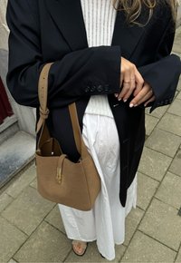 Bolsa de mão em couro sintético castanho com forma estruturada e fecho dourado. Usada com um blazer preto sobre um top de malha branco e saia branca.