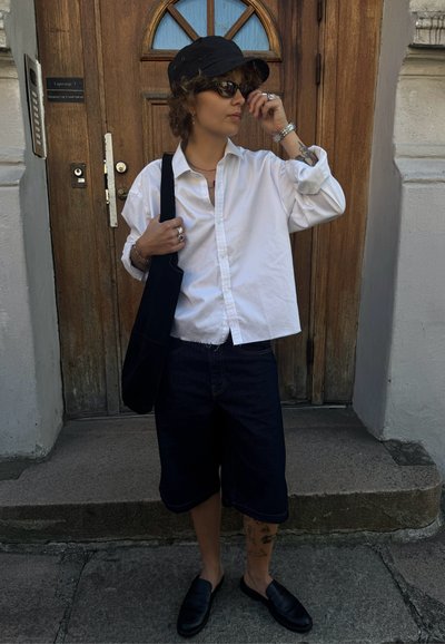 Camisa blanca de botones, pantalones cortos de mezclilla oscuros, gorra negra, gafas de sol y zapatos negros sin cordones. Los accesorios incluyen anillos y un bolso de hombro.