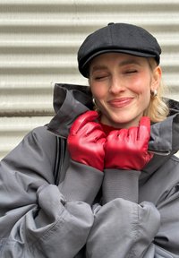 Guanti rossi in pelle in posizione chiusa, indossati con una giacca grigia oversize. Completa il look un berretto nero e orecchini a cerchio in argento.