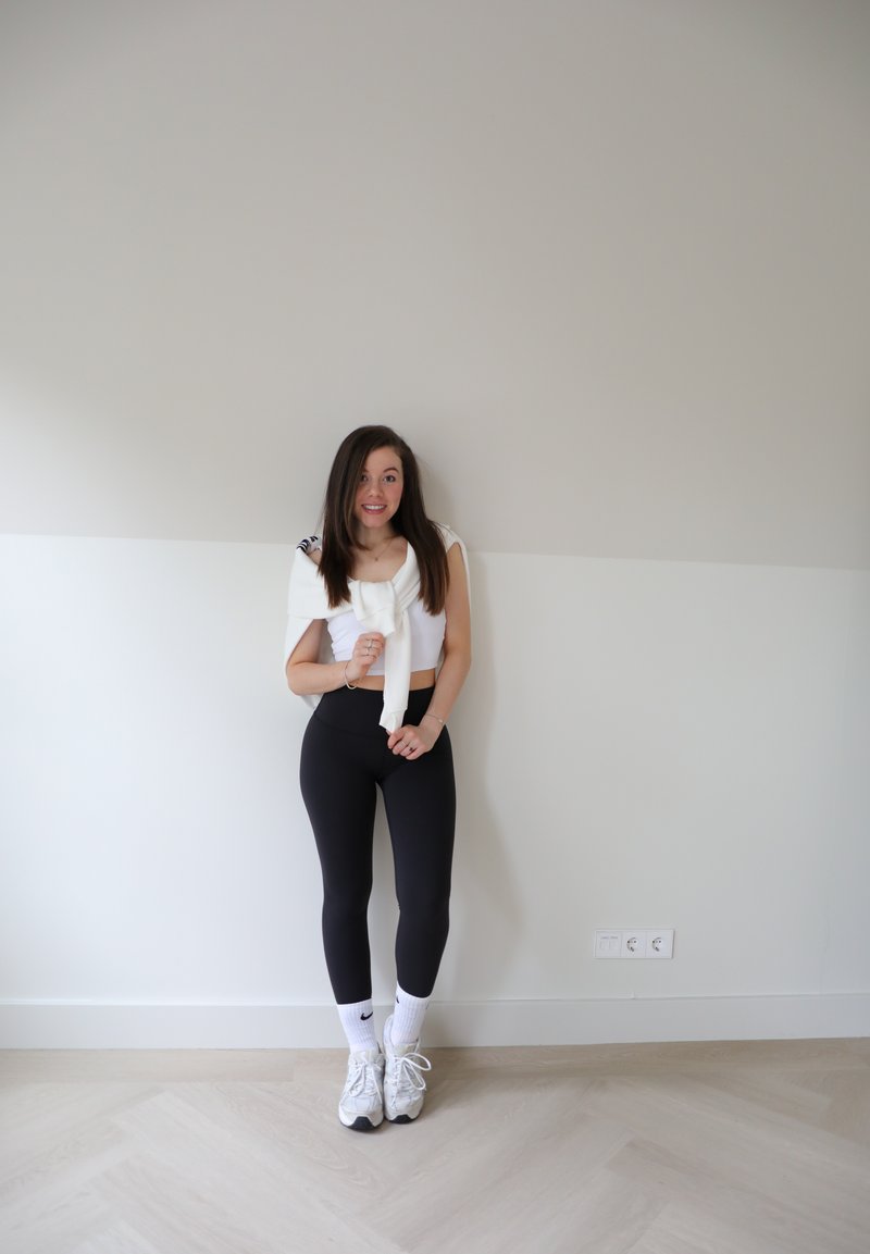 Άσπρο crop top με λεπτομέρεια φιόγκος, μαύρες κολάν, άσπρα παπούτσια Nike, και άσπρες κάλτσες, στέκεται μπροστά σε έναν ανοιχτό γκρίζο τοίχο.