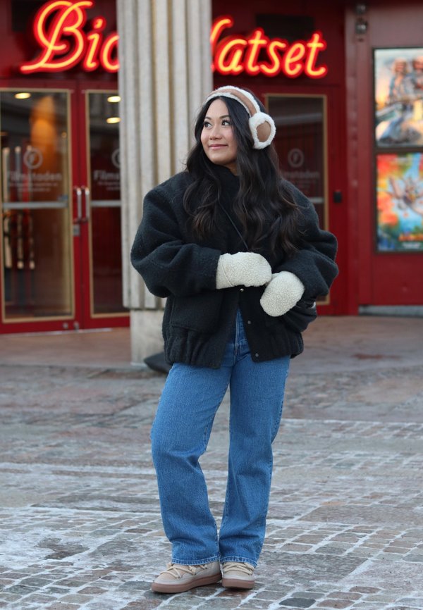 Zwarte, donzige oversized jas, lichtgekleurde wanten, blauwe spijkerbroek en beige schoenen. Lang, donker haar en oorwarmers. Vastgelegd in een stedelijke omgeving.