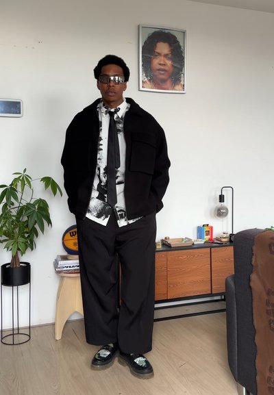 Chaqueta negra de gran tamaño, camisa blanca con patrón abstracto negro, corbata negra, pantalones negros holgados, zapatos negros y verdes, ambiente interior con planta.