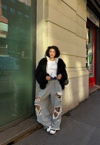 Svart jakke med kunstpels, hvit tettsittende topp, grå revne joggebukser med frynsete kanter, og hvite slip-on sko. Står ved et vindu i glass.