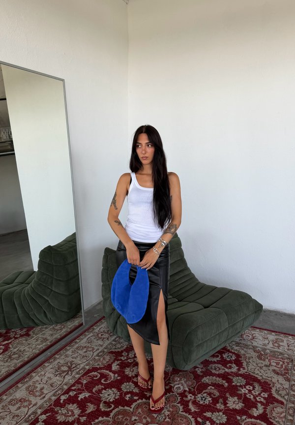 Femme aux longs cheveux foncés portant un débardeur blanc et une jupe noire fendue, tenant un sac bleu, debout près d'une chaise verte rembourrée et d'un grand miroir.