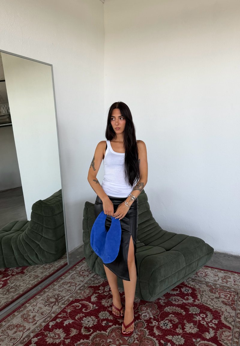 Femme aux longs cheveux foncés portant un débardeur blanc et une jupe noire fendue, tenant un sac bleu, debout près d'une chaise verte rembourrée et d'un grand miroir.