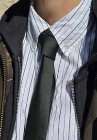 White button-down shirt with black vertical stripes, paired with a solid black tie. Encased in a zippered dark jacket.