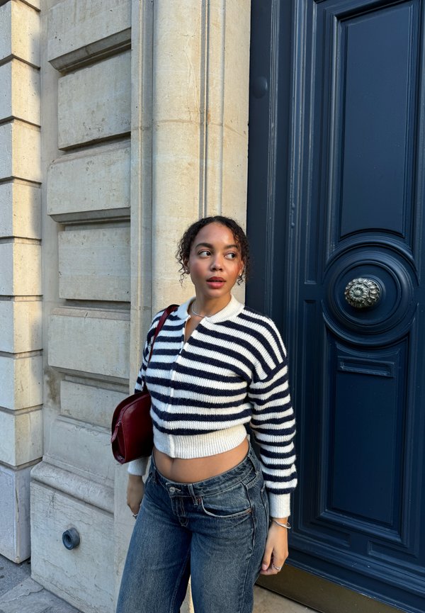 Gestreifter, kurz geschnittener Pullover in Marineblau und Weiß, kombiniert mit hochtaillierten Jeans. Rote Handtasche und strukturierte Backsteinmauer im Hintergrund.