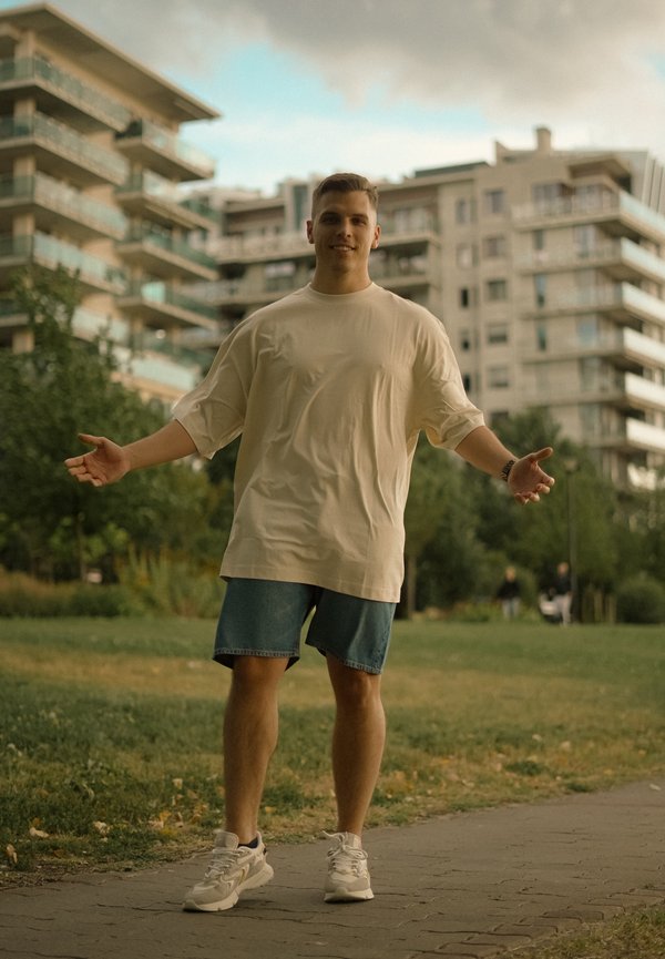 Ung mand iført beige oversized t-shirt og denimshorts står på en sti med åbne arme i en bypark med moderne bygninger bagved.