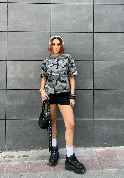 Camiseta oversized de camuflaje gris, pantalones cortos negros, zapatillas negras gruesas, calcetines blancos, accesorios plateados y una bolsa negra con acentos metálicos.