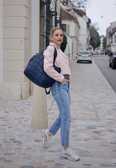 Mochila acolchada azul marino, sudadera corta rosa claro, jeans azul de cintura alta y zapatillas blancas con detalles negros, caminando sobre pavimento de piedra.