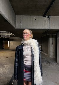 Jeune femme en manteau en cuir noir et écharpe blanche duveteuse, debout dans un parking souterrain en béton sombre, les yeux fermés, tournée vers la gauche.