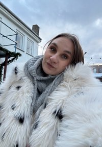 Femme en plein air portant un manteau en fourrure blanc et une écharpe grise, avec un bâtiment et un ciel nuageux en arrière-plan.