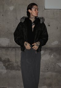 Črna usnjena jakna z ovratnikom obloženim z dlako, črna majica in sive hlače z navpičnimi črtami. Tetovaže so vidne na rokah in vratu. Enostavno ozadje.