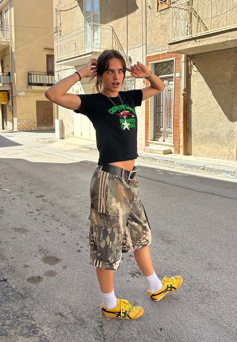 Schwarzes kurzes T-Shirt mit grüner Schrift, camouflierte Cargo-Shorts, gelbe Sneakers, weiße Socken, stehend auf einer Straße vor einer beigen Wand.