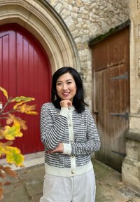 Grå og hvitstripet cardigan med perleknapper, båret over en mørk topp; lyse bukser; satt mot en steinmur og rød dør.