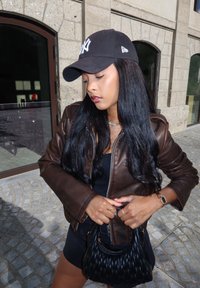 Καφέ δερμάτινο μπουφάν, μαύρο καπέλο μπέιζμπολ με λογότυπο, μαύρη μπλούζα, μαύρη πλεκτή τσάντα, μακριά σκούρα μαλλιά, στέκεται σε επιφάνεια από πλακόστρωτο.