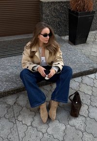 Beige, dupla soros kabát szürke felső felett, kék trapéz farmernadrág, bézs velúr bokacsizma, fekete kézitáska, fekete napszemüveg, kőből készült burkolaton ülve.