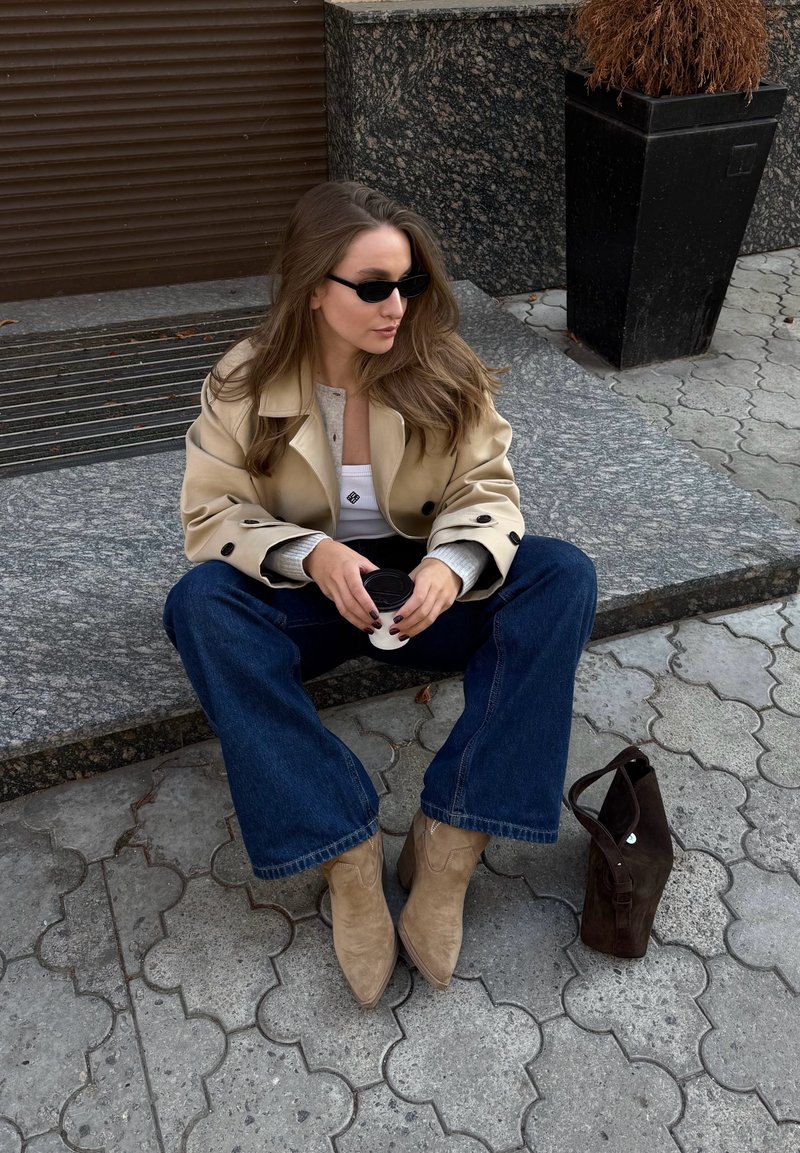 Beiger dreireihiger Mantel über grauem Oberteil, blaue Schlagjeans, tanfarbene Wildleder-Stiefeletten, schwarze Handtasche, schwarze Sonnenbrille, sitzend auf Steinpflaster.
