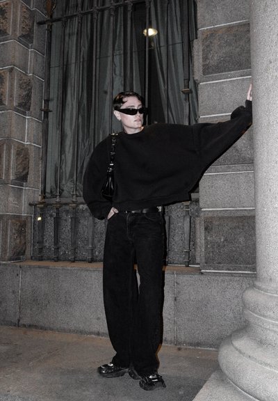 Sudadera negra de gran tamaño, jeans negros y zapatos negros gruesos. Los accesorios incluyen un bolso negro y gafas de sol oscuras. Fondo de piedra.