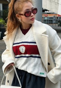 Palton din lână albă peste un pulover tricotat roșu și alb cu dungi albastre și un logo Adidas. Pantaloni subțiri cu dungi bleumarin și o geantă albă.