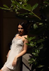 Φόρεμα σε ανοιχτό μπεζ χρώμα με χαλαρή γραμμή, δέσιμο μπροστά με κορδόνια και λεπτές τιράντες, συνοδευόμενο από μπλε τσάντα. Πράσινα φύλλα πλαισιώνουν τη σύνθεση.