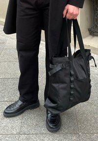 Melna liela soma no izturīga materiāla ar regulējamām siksnām, sānu kabatām un gludu tekstūru. Valkāta kopā ar melniem apaviem un biksēm.