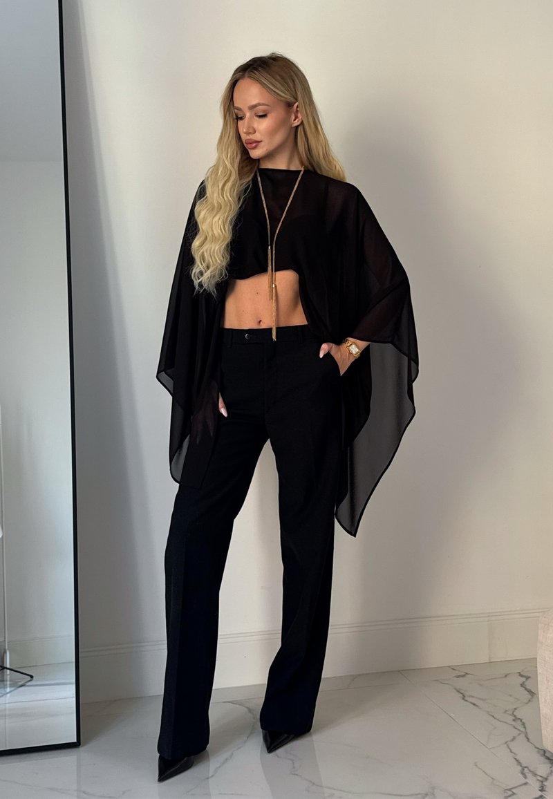 Blond žena nosi čierny priehľadný pončo, krátky top, nohavice so širokými nohavicami a špicaté opätky, stojí vo vnútri na mramorovej podlahe s jednou rukou v vrecku.