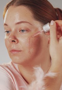 Kvinne med rødt hår påfører rosa ansiktsserum med en dråpeteller, lar væsken renne nedover venstre kinn med et nøytralt uttrykk.