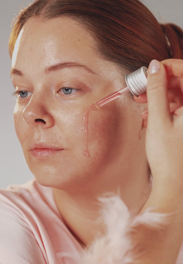 Kvinna med rött hår applicerar rosa ansiktsserum med en pipett, låter vätskan droppa ner längs hennes vänstra kind med ett neutralt uttryck.