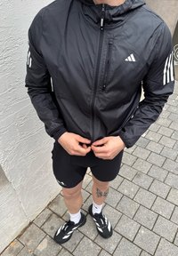 Personne portant une veste de course Adidas noire et un short, en train de fermer la fermeture éclair de la veste, debout sur un trottoir avec un tatouage visible sur la jambe et des baskets noires.