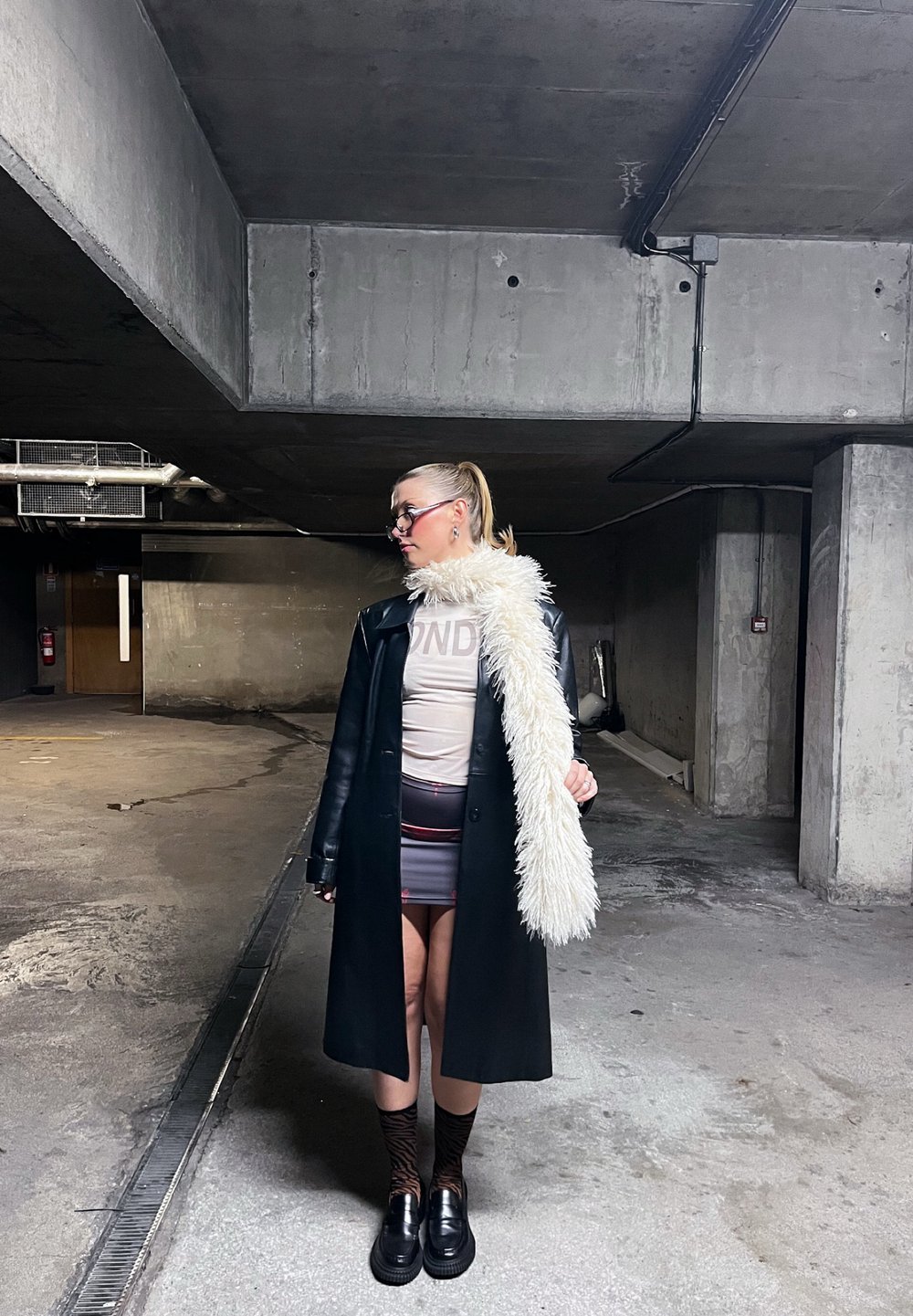 Femme avec queue de cheval portant des lunettes, un long manteau noir, une écharpe blanche en fourrure, une jupe imprimée et des chaussures à plateforme, se tenant dans un parking en béton faiblement éclairé.