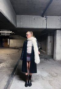 Femme avec queue de cheval portant des lunettes, un long manteau noir, une écharpe blanche en fourrure, une jupe imprimée et des chaussures à plateforme, se tenant dans un parking en béton faiblement éclairé.