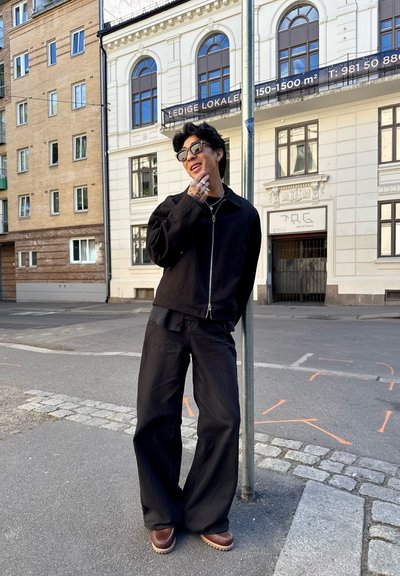 Chaqueta negra con cremallera sobre una camisa gris, combinada con pantalones anchos negros y zapatos de plataforma marrones. Entorno urbano con edificios de ladrillo y blancos.