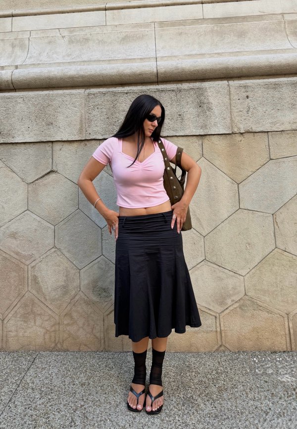 Vrouw met lang donker haar, die een roze top, zwarte midirok, zwarte sokken met sandalen en een zonnebril draagt, staat tegen een zeshoekige stenen muur.