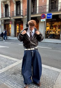 Brązowa skórzana kurtka typu bomber na białej koszuli, kratkowany krawat i oversize'owe niebieskie dżinsy z srebrnymi guzikami, stojące na brukowanej ulicy.