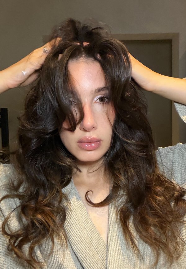 Donna con capelli lunghi, mossi e scuri che indossa un maglione beige e tiene i capelli sollevati con entrambe le mani, guardando direttamente la fotocamera al chiuso.