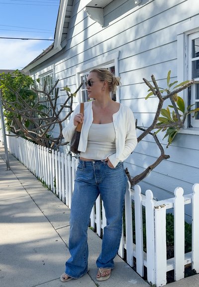 Mujer con cárdigan blanco, camiseta sin mangas, vaqueros azules y sandalias, de pie junto a una cerca blanca de estacas con ramas de árbol sin hojas cerca de una casa.
