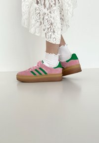 Sneakers di suede rosa con accenti verdi, tre righe sui lati e una suola spessa in gomma, abbinate a calzini bianchi con balze e una gonna in pizzo.