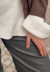 Pull en maille côtelée blanche avec larges poignets, superposé sous une veste marron. Pantalon en cuir noir visible, avec une main reposant sur la hanche.