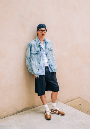 Denim jacket, blauwgestreept shirt, zwarte streepjes korte broek, en bruine sneakers. Staand tegen een beige muur, met witte sokken.