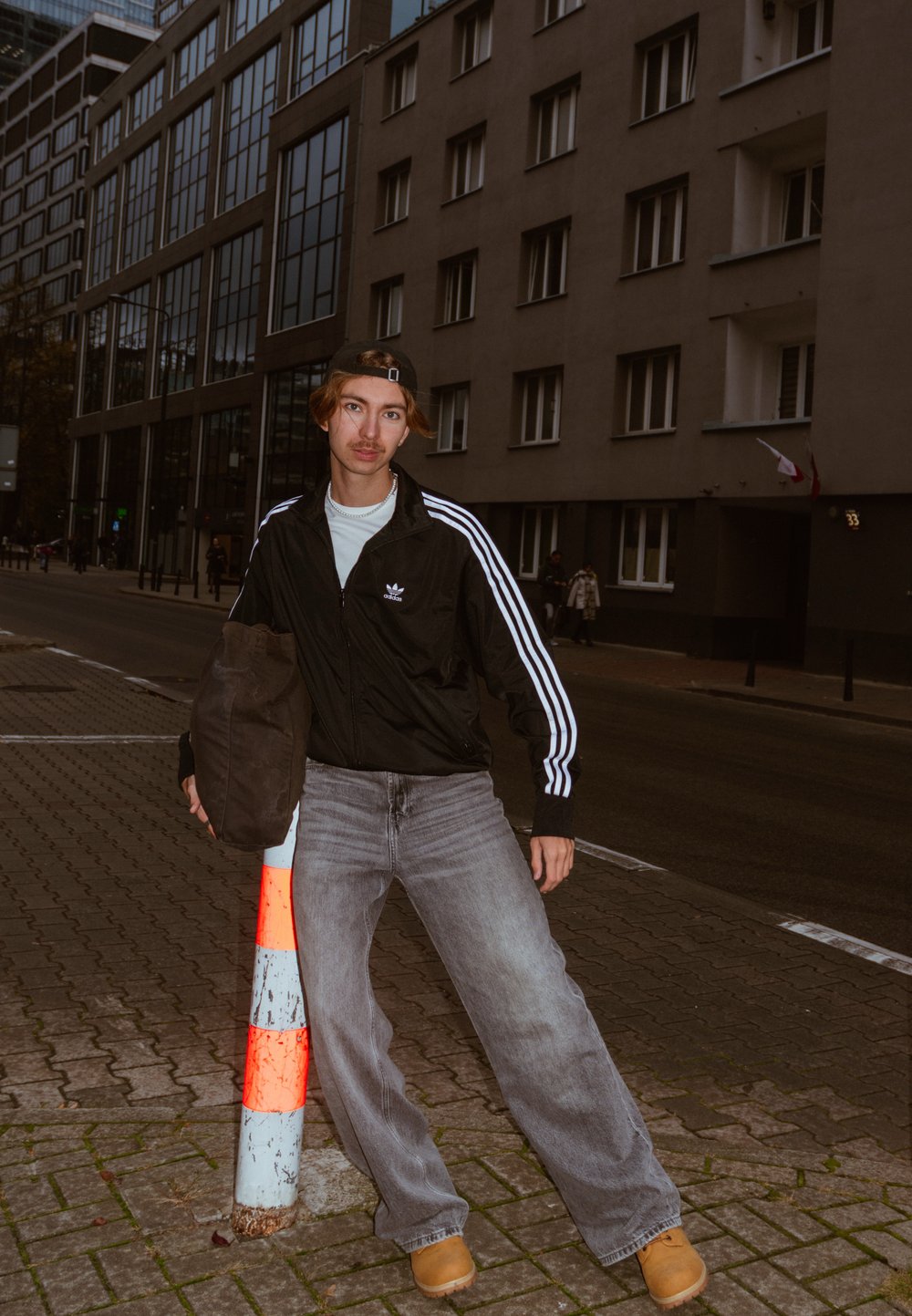 Jauns vīrietis melnā Adidas jaku un pelēkās džinsās, atbalstoties uz baltas un oranžas svītrainas staba pilsētas ietves pievakarē.