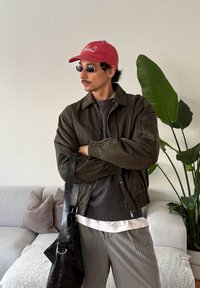 Άνδρας με κόκκινο καπέλο, γυαλιά ηλίου, καφέ μπουφάν, γκρι πουλόβερ και ριγέ παντελόνι, κρατώντας μια μαύρη δερμάτινη τσάντα σε εσωτερικό χώρο κοντά σε φυτό και καναπέ.