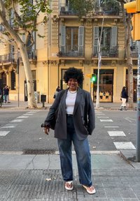 Čierne sako cez bielu košeľu, široké modré rifle, žabky a textúrovaný afro účes; stojí na mestskej ulici s budovami v pozadí.