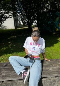 Weißes Nike-Grafik-T-Shirt, hellblaue Jeans, pink akzentuierte schwarze Sneakers, silberner Gürtel mit Kette, hält ein Smartphone und sitzt auf einer Bank.
