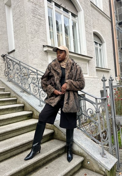 Abrigo de piel marrón con cuello tipo chal, camiseta negra y pantalones cortos negros de pierna ancha. Botas negras hasta la rodilla con punta puntiaguda. Gorra y pañuelo envolvente en la cabeza.